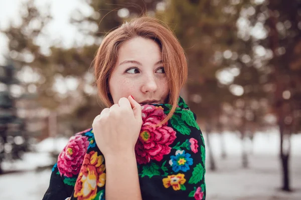 Russian beauty woman with red hair and freckles in the national scarf stands on a background of trees. — Stock Photo, Image