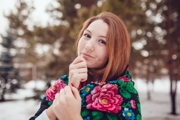 Russische Schönheit mit roten Haaren und Sommersprossen im Nationalschal steht auf einem Baumhintergrund. — Stockfoto