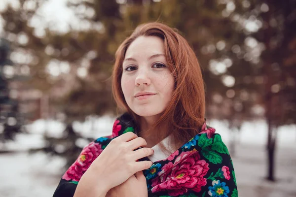 Russian beauty woman with red hair and freckles in the national scarf stands on a background of trees. — Stock Photo, Image