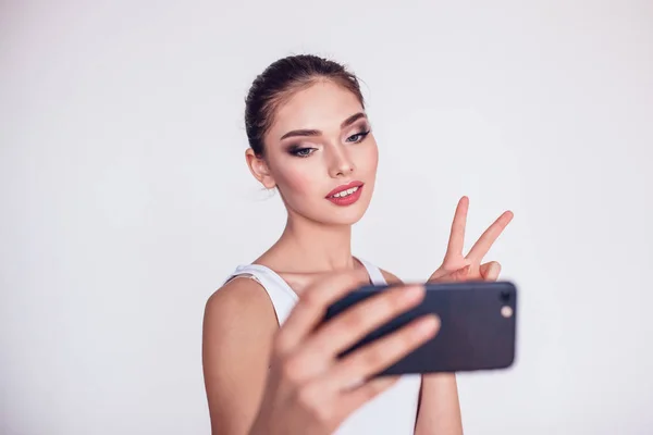 Όμορφη μελαχρινή κοπέλα με μακιγιάζ κάνει selfie σε λευκό φόντο. — Φωτογραφία Αρχείου