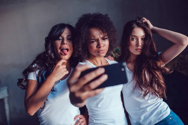 Három gyönyörű mosolygó lány a fehér póló grimaszol, és hogy selfie használ smartphone. — Stock Fotó