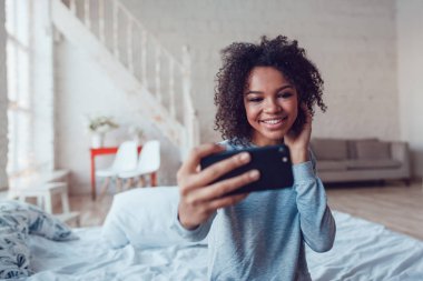 Çok gülümseyen Afrikalı kız yatakta evde oturan selfie yapmak.