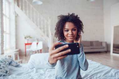 Çok gülümseyen Afrikalı kız yatakta evde oturan selfie yapmak.