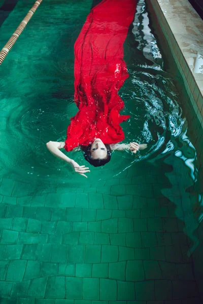 Mujer morena de moda con hermoso maquillaje profesional flotando en una piscina de tela roja . —  Fotos de Stock