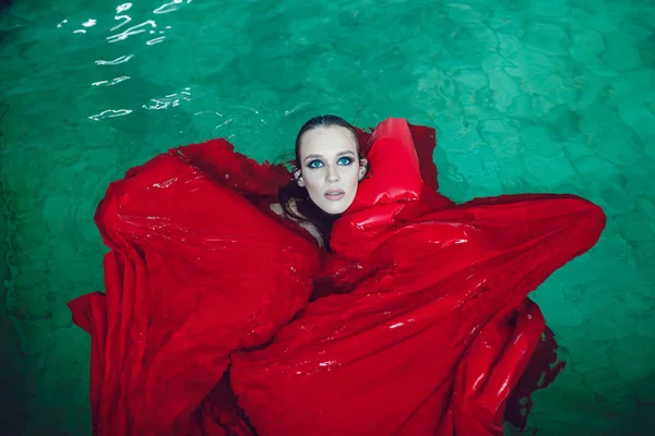 Mujer morena de moda con hermoso maquillaje profesional flotando en una piscina de tela roja . — Foto de Stock