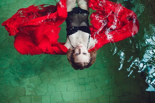 Femme de mode avec belle maquillage professionnel flottant dans une piscine de tissu rouge . — Photo