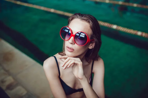 Mulher morena bonita em óculos de sol engraçados com um profissional compõem sentado perto da piscina . — Fotografia de Stock