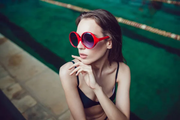 Hermosa mujer morena en gafas de sol divertidas con un maquillaje profesional sentado cerca de la piscina . — Foto de Stock