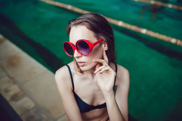 Hermosa mujer morena en gafas de sol divertidas con un maquillaje profesional sentado cerca de la piscina . — Foto de Stock