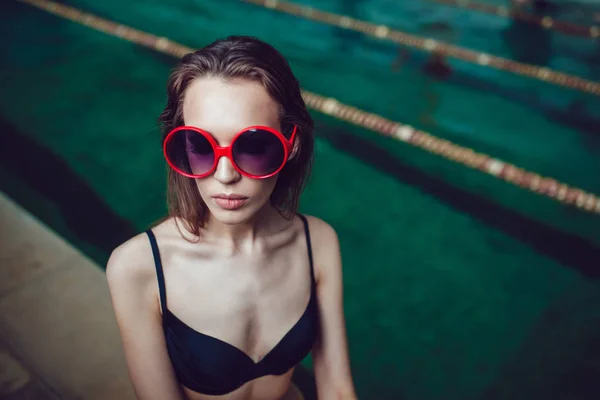 Beautiful brunette woman in funny sunglasses with a professional make up sitting near the pool. — Stock Photo, Image
