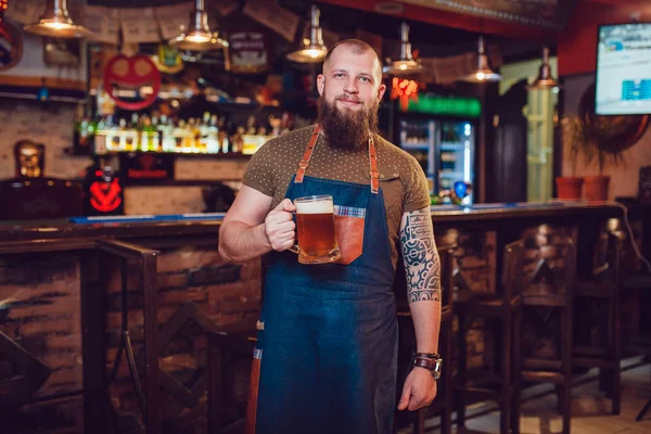 Bearded barman with tattoos wearing an apron standing near the bar and holding a glass of beer — Stock Photo, Image