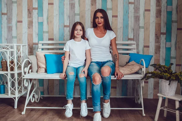 Mamá e hija con el pelo largo hermoso en ropa idéntica están sentadas en un sofá de madera. Camiseta blanca en blanco . — Foto de Stock