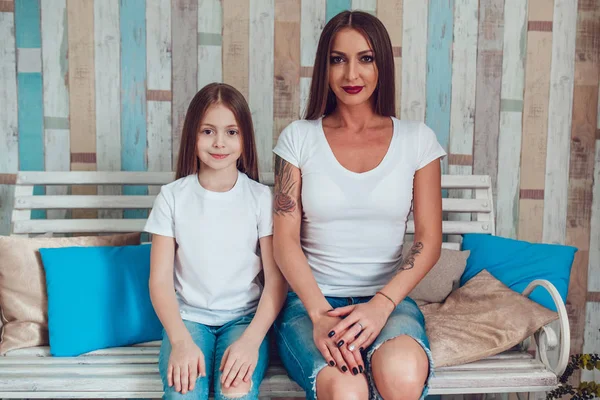 Mamá e hija con el pelo largo hermoso en ropa idéntica están sentadas en un sofá de madera. Camiseta blanca en blanco . — Foto de Stock