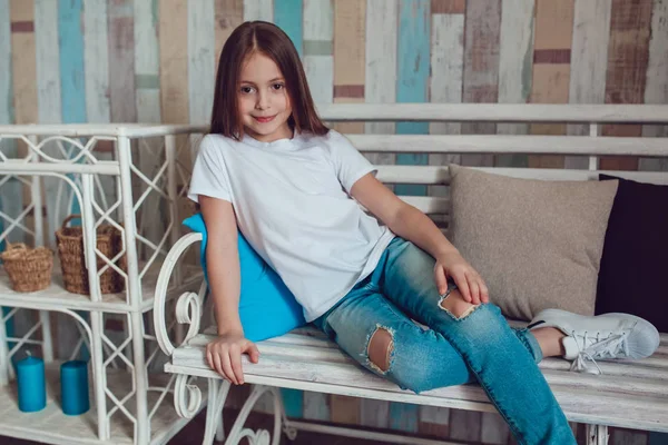 Śliczną dziewczynkę w pusty biały T-shirt i dżinsy siedzi na kanapie drewniane. — Zdjęcie stockowe