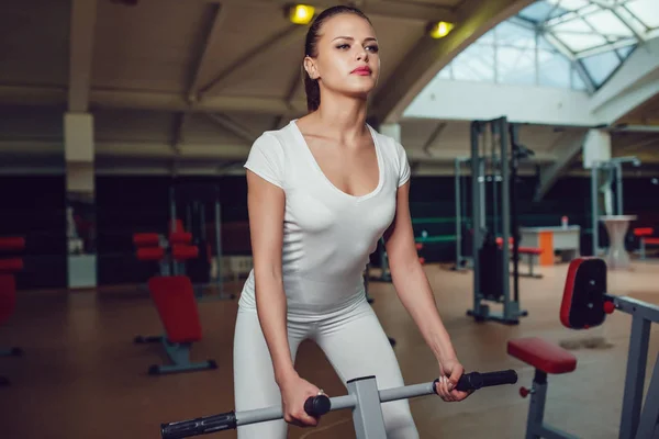 Hermosa chica haciendo ejercicios en el simulador en el gimnasio vestido en blanco camiseta blanca y medias. Prepárate. . — Foto de Stock