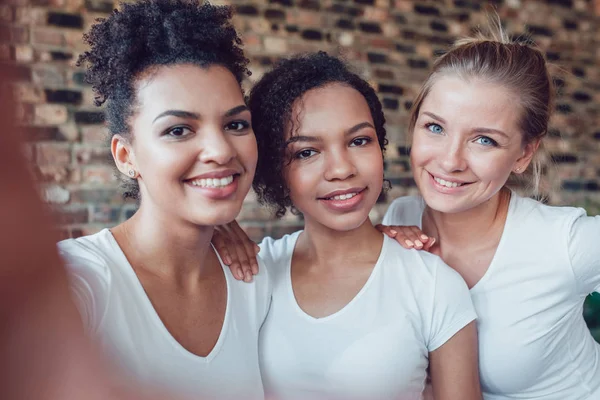 Mooie en grappige meisjes in witte T-shirts maakt selfie. Twee afro-Amerikanen en een Europese. — Stockfoto