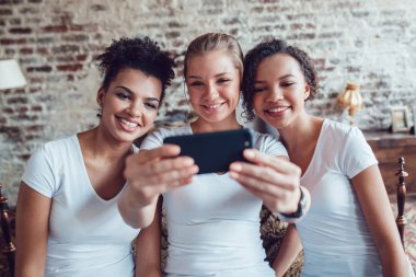 Güzel kızlar eğleniyor ve yatağın oturan selfie olun. İki afro-Amerikalı ve Avrupalı kız.