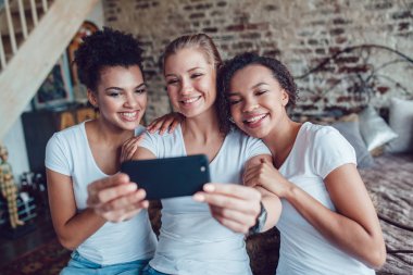 Güzel kızlar eğleniyor ve yatağın oturan selfie olun. İki afro-Amerikalı ve Avrupalı kız.