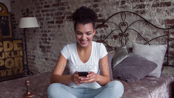 Mulher afro-americana sorridente atraente usando smartphone sentado na cama . — Vídeo de Stock