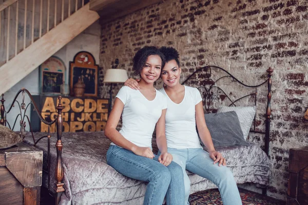 Dos chicas afroamericanas atractivas en camisetas blancas están sentadas en la cama . — Foto de Stock