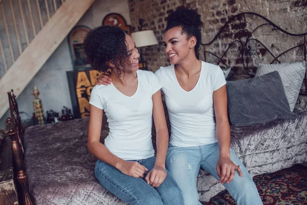Dos chicas afroamericanas atractivas en camisetas blancas están sentadas en la cama . — Foto de Stock