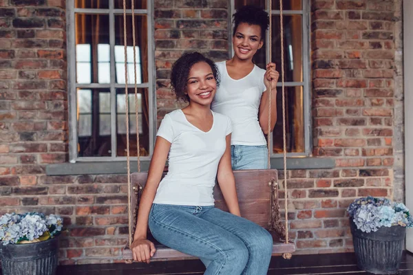 Aantrekkelijke Afrikaans Amerikaans meisje in witte T-shirts zittend op een schommel op een bakstenen muur achtergrond. Het tweede meisje staat in de buurt. — Stockfoto