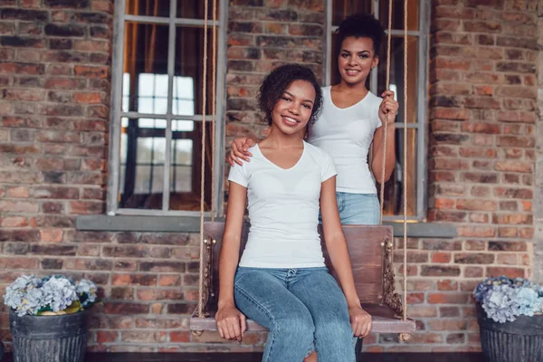 Atractiva chica afroamericana en camisetas blancas sentada en un columpio sobre un fondo de pared de ladrillo. La segunda chica está de pie cerca . —  Fotos de Stock