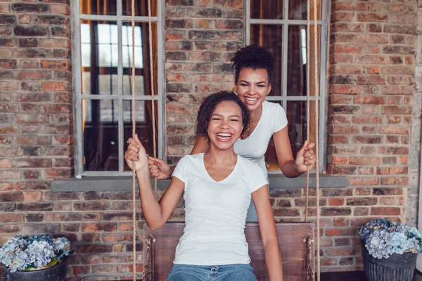 Dwie atrakcyjne african american dziewczyny w białych koszulkach, kołysanie na huśtawce na tle ściany Cegła. — Zdjęcie stockowe