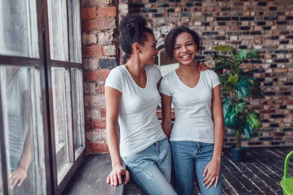 Dwie atrakcyjne african american dziewczyny w białych koszulkach stoją w pobliżu okna. — Zdjęcie stockowe