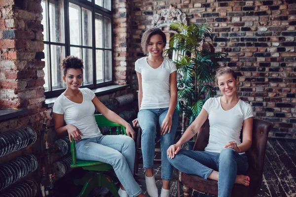 Tres chicas atractivas en camisetas blancas están sentadas en sillones cerca de la ventana . — Foto de Stock