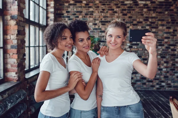 Tres chicas atractivas en camisetas blancas hacen selfie cerca de la ventana . — Foto de Stock