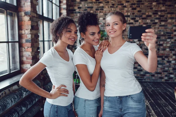 Tres chicas atractivas en camisetas blancas hacen selfie cerca de la ventana . — Foto de Stock