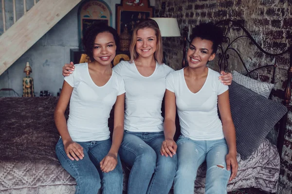 Trzy atrakcyjne dziewczyny w białych koszulkach siedzą w domu na łóżku. — Zdjęcie stockowe