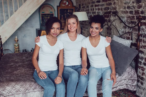 Tres chicas atractivas en camisetas blancas están sentadas en casa en la cama . — Foto de Stock