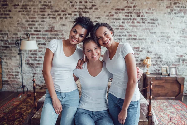 Trzy atrakcyjne dziewczyny w białych koszulkach siedzą w domu koło łóżka. — Zdjęcie stockowe