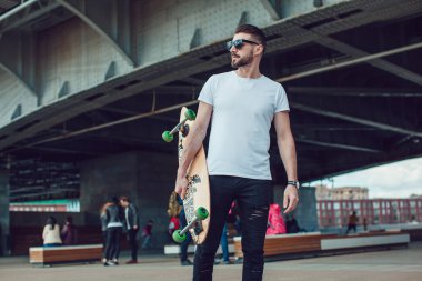 Şık erkek güneş gözlüğü ve sakalı ile uzun bir tahta ile sokakta duruyor. T-shirt alay.