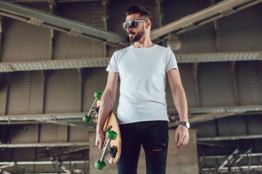 Şık erkek güneş gözlüğü ve sakalı ile uzun bir tahta ile sokakta duruyor. T-shirt alay.