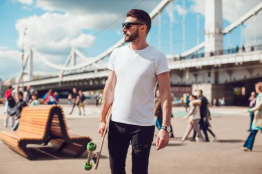 Şık erkek güneş gözlüğü ve sakalı ile uzun bir tahta ile sokakta duruyor. T-shirt alay.