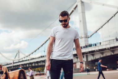 Şık erkek güneş gözlüğü ve sakalı ile uzun bir tahta ile sokakta duruyor. T-shirt alay.