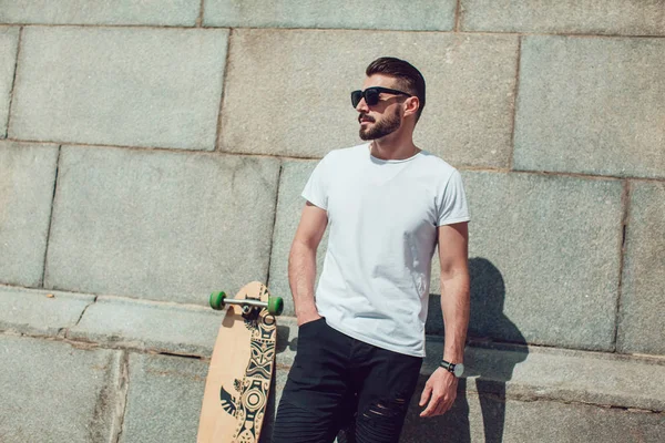 Stylish man in sunglasses and with a beard stands on wall background with a long board. T-shirt mock up.