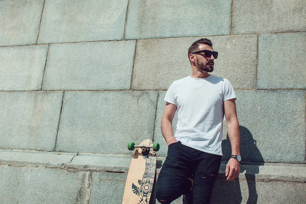 Stylish man in sunglasses and with a beard stands on wall background with a long board. T-shirt mock up.