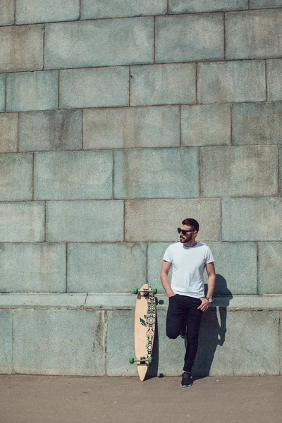 Stylish man in sunglasses and with a beard stands on wall background with a long board. T-shirt mock up.