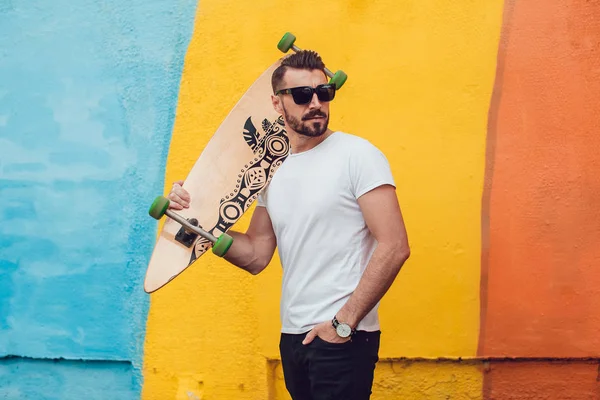 Stylish man in sunglasses and with a beard stands on the background of a multi-colored wall with a long board. T-shirt mock up.