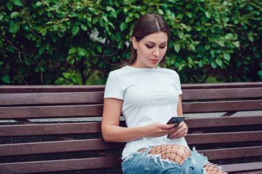 Çekici kız beyaz park ve smartphone kullanarak bir bankta oturuyor.