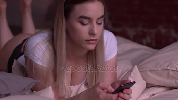 Сексуальная блондинка лежит на кровати и пользуется смартфоном . — стоковое видео