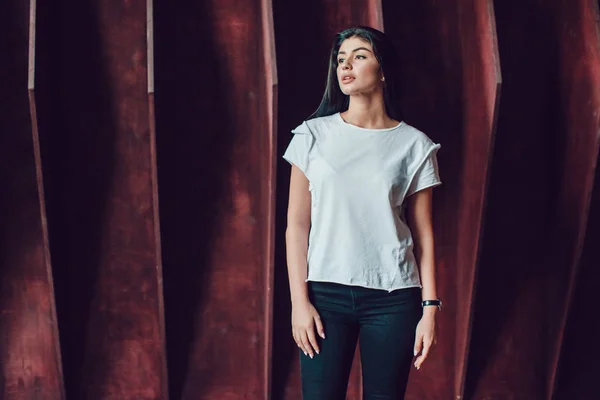 Die attraktive brünette Frau in weißem T-Shirt steht vor dunklem Holzwandhintergrund. Attrappe. — Stockfoto