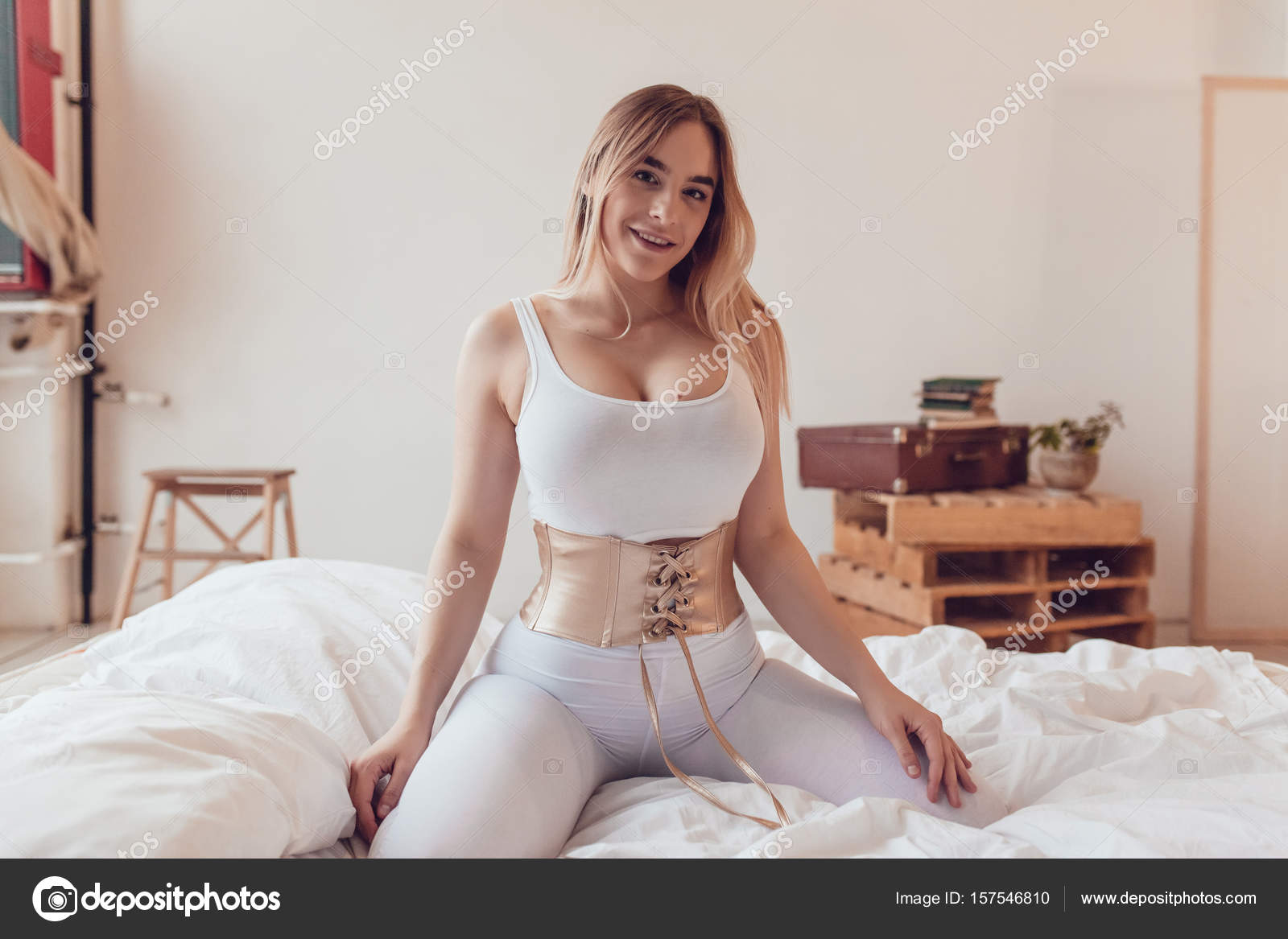 Sexy blonde woman with big breasts in a white top and corset sits on the bed.  Stock Photo by ©airkost 157546810