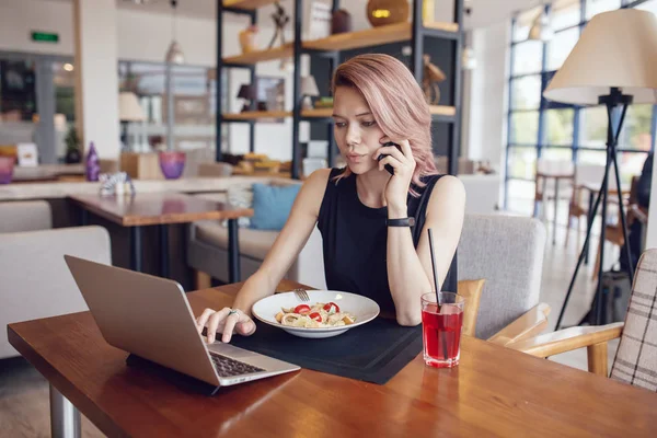 Mujer de negocios come en un café y utiliza un teléfono inteligente y un ordenador portátil . — Foto de Stock