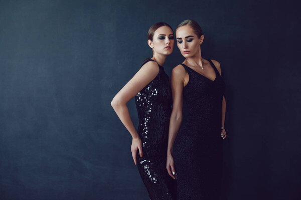 Two beautiful women in black night fashion dress posing on a black background.