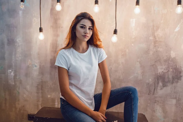 Sexy vrouw in een witte T-shirt op een grijze achtergrond met lampen. Mock-up. — Stockfoto